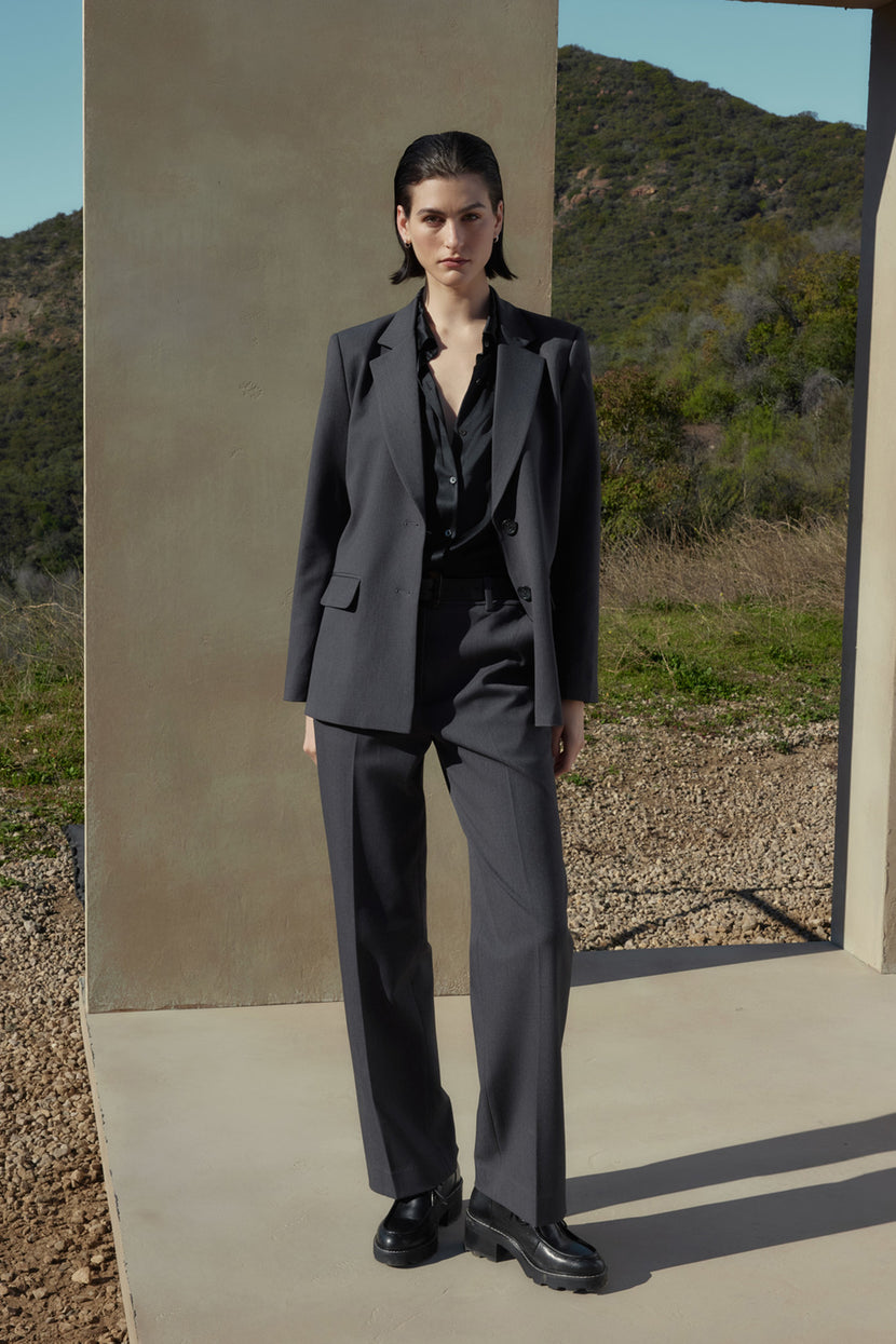 A person with short dark hair wears a grey suit made of suiting fabric and a black shirt, standing on a concrete platform with a mountainous landscape in the background. The PRINCE PANT from Velvet by Jenny Graham completes the polished look.