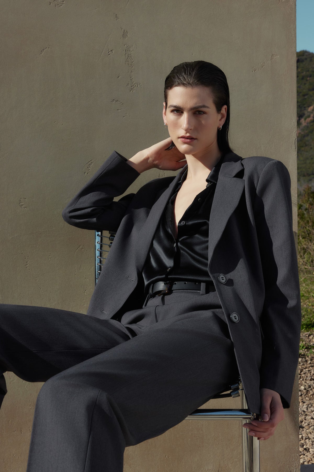   A person wearing a dark grey suit and black shirt, featuring the PRINCE PANT straight leg trousers in suiting fabric by Velvet by Jenny Graham, sits on a chair against a neutral background, with one hand resting behind their neck. 