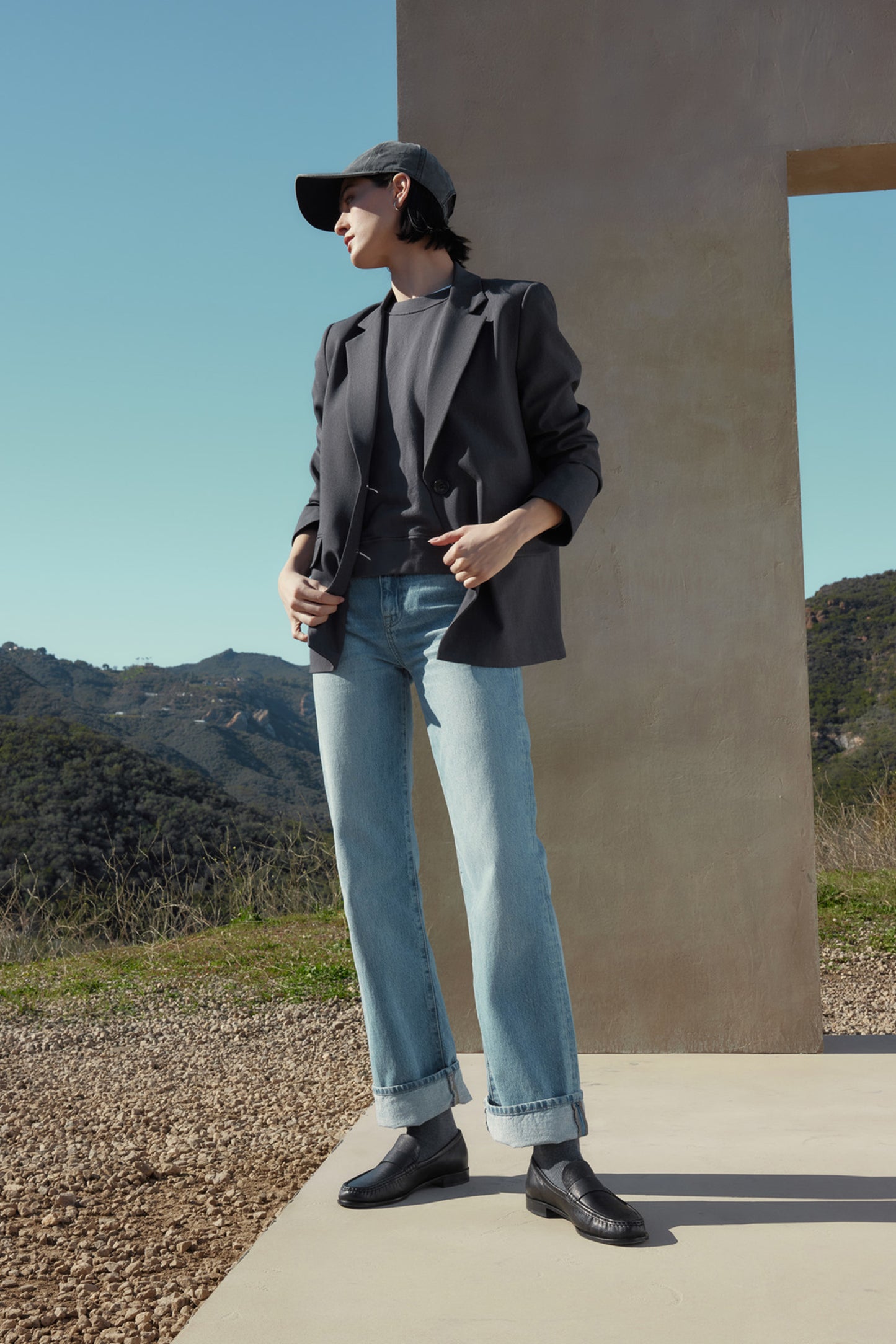 A person wearing a black cap, dark blazer, YNEZ SWEATSHIRT by Velvet by Jenny Graham made of organic cotton, and light blue jeans stands near a structure with a hilly landscape in the background.-37666967126209