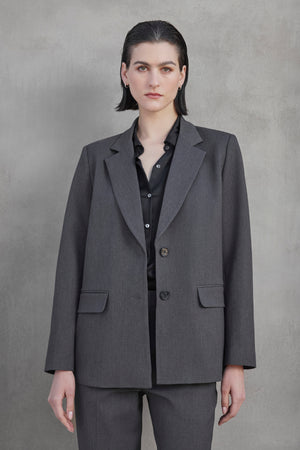 Individual with short hair clad in a REXFORD BLAZER by Velvet by Jenny Graham, paired with a black shirt, standing against a plain gray background.