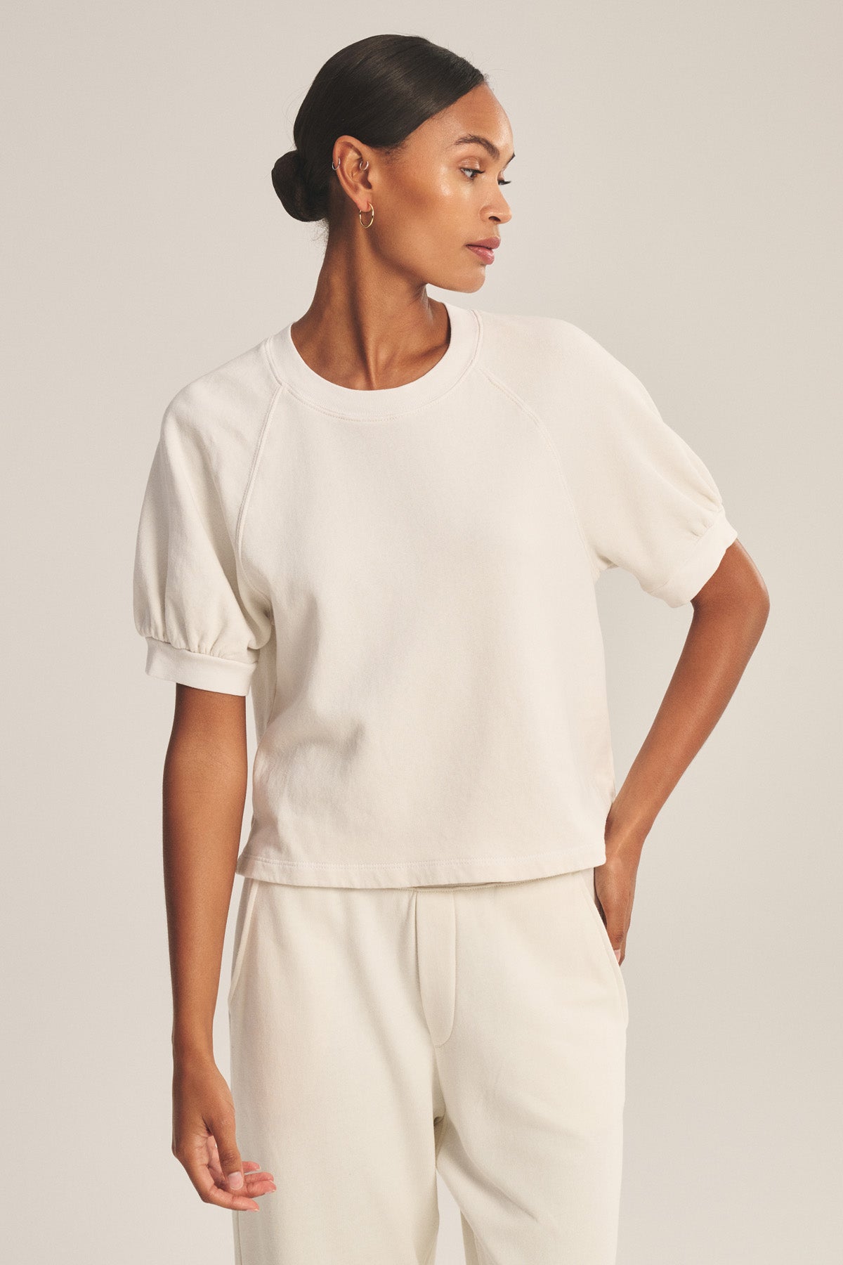 A woman wears the SONNET SWEATSHIRT by Velvet by Graham & Spencer—a cream-colored, cotton fleece boxy cropped top with matching pants—standing against a plain background, looking left with one hand on her hip.-41044145897665