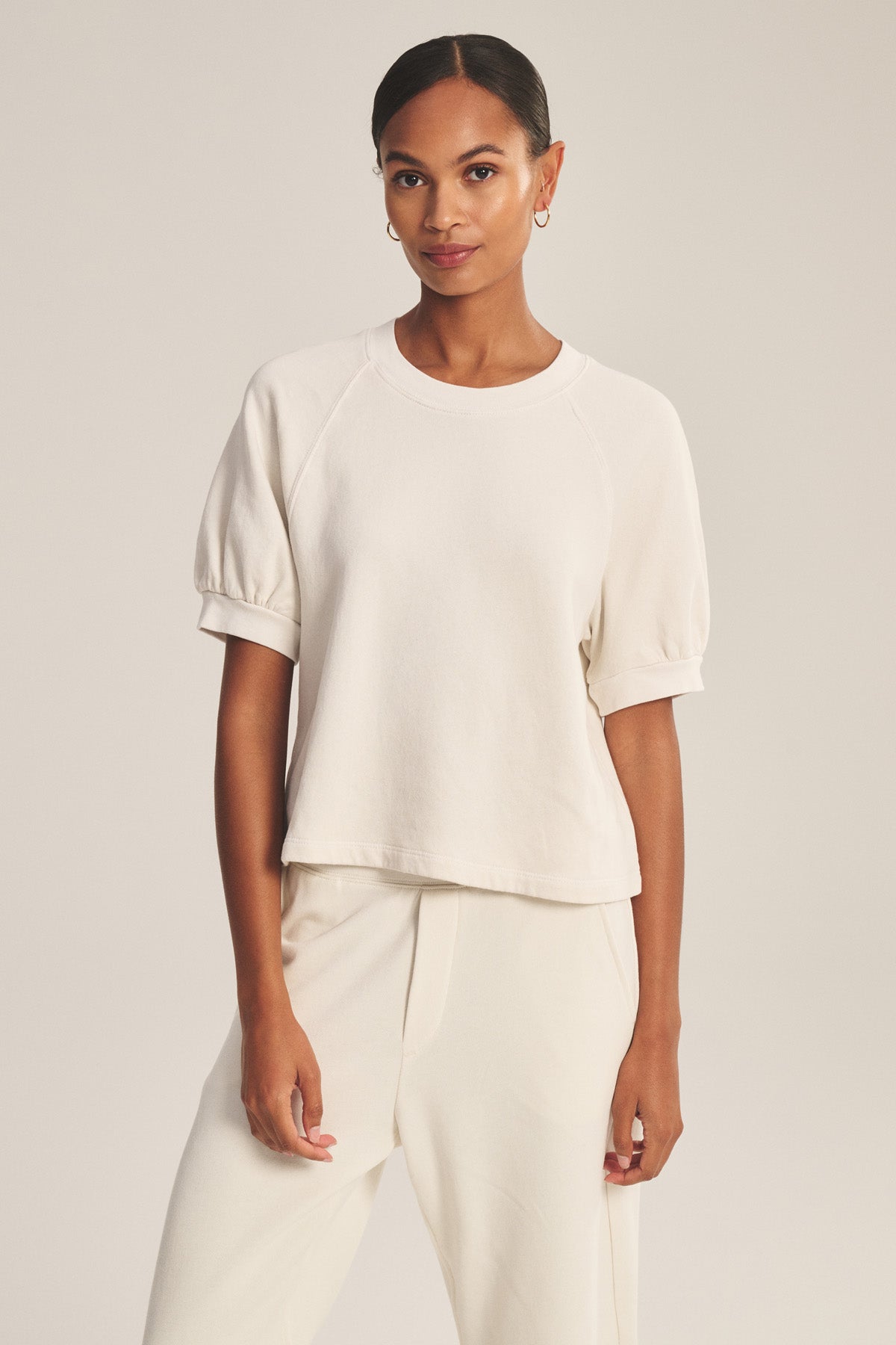 A woman faces forward wearing the Velvet by Graham & Spencer SONNET SWEATSHIRT in cream, styled with matching pants, her hair pulled back and minimal accessories.-41044145864897