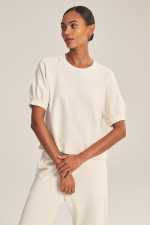 A woman stands against a plain background wearing the off-white SONNET SWEATSHIRT with puff raglan sleeves by Velvet by Graham & Spencer, paired with matching off-white pants.