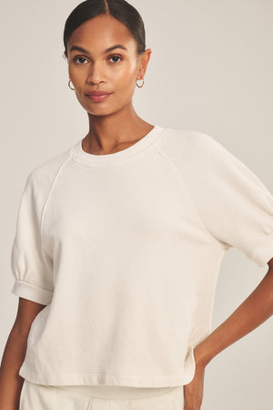 A woman poses with one hand on her hip, wearing the SONNET SWEATSHIRT by Velvet by Graham & Spencer—an off-white cotton fleece top with short puff raglan sleeves—against a neutral background.