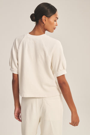 A woman stands facing away, wearing the off-white SONNET SWEATSHIRT by Velvet by Graham & Spencer with matching pants. Her hair is tied back in a low bun.