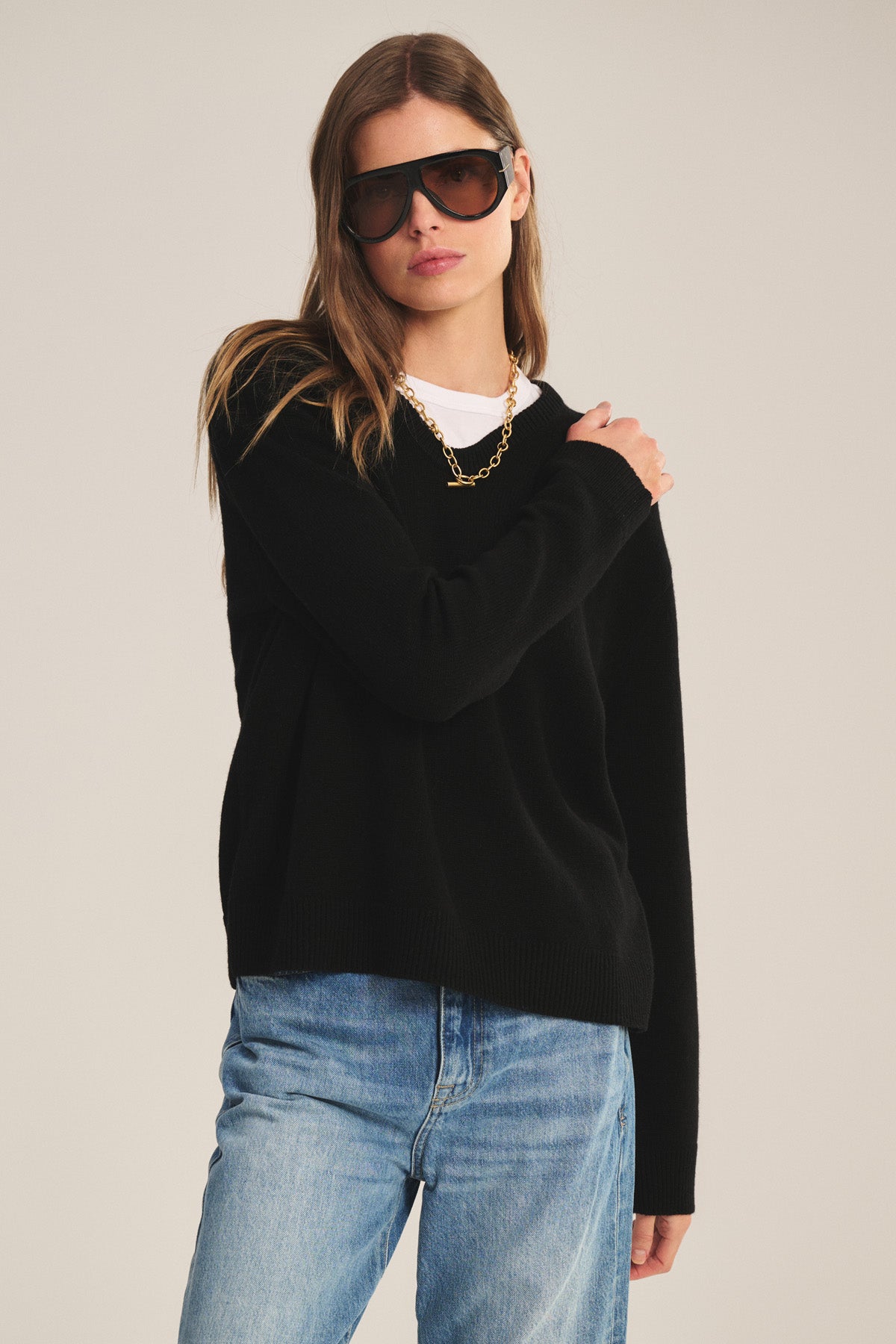 A woman in Velvet by Graham & Spencer's SORREL SWEATER—a black cashmere-wool knit with a U-neck—layered over a white shirt, blue jeans, and gold chain necklace, stands against a plain background.-40808702673089