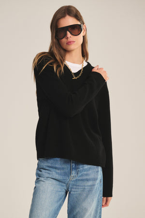 A woman in Velvet by Graham & Spencer's SORREL SWEATER—a black cashmere-wool knit with a U-neck—layered over a white shirt, blue jeans, and gold chain necklace, stands against a plain background.