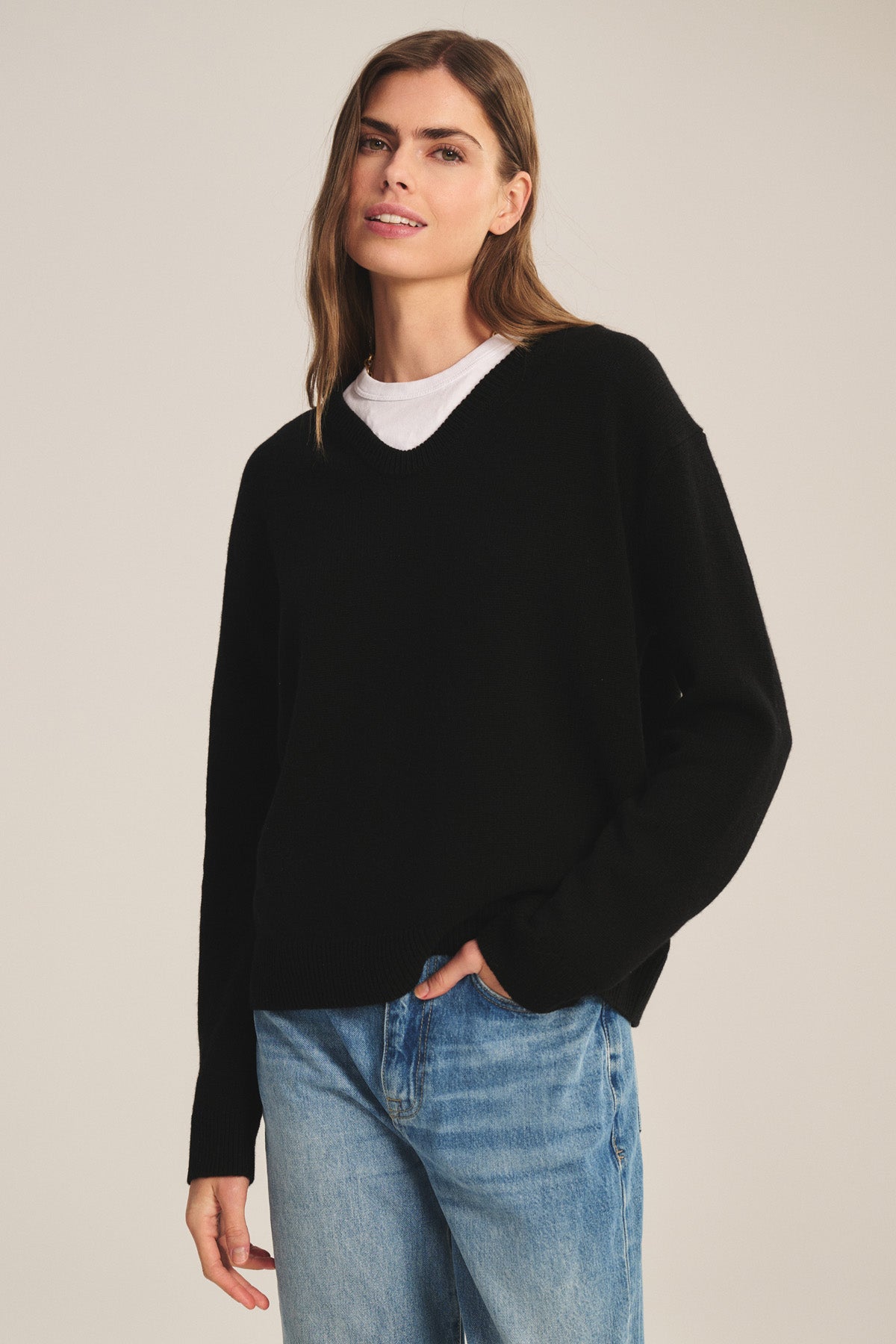 A woman wears the SORREL SWEATER by Velvet by Graham & Spencer, a black cashmere-wool piece with a U-neckline, layered over a white T-shirt and blue jeans, standing against a plain light background.