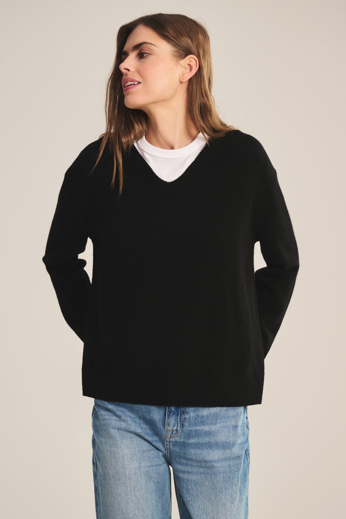 A person wears the SORREL SWEATER by Velvet by Graham & Spencer—a black cashmere-wool knit with a modern silhouette—layered over a white T-shirt and blue jeans, standing hands behind back against a neutral background.-40808702509249