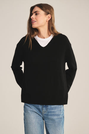 A person wears the SORREL SWEATER by Velvet by Graham & Spencer—a black cashmere-wool knit with a modern silhouette—layered over a white T-shirt and blue jeans, standing hands behind back against a neutral background.