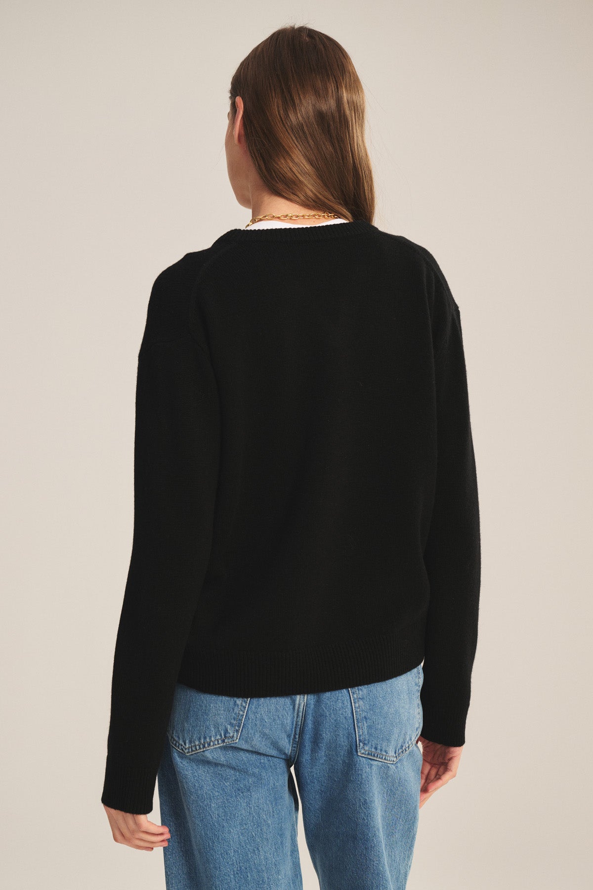 A person wearing the SORREL SWEATER by Velvet by Graham & Spencer, a black cashmere-wool blend with a modern silhouette, and blue jeans stands facing away from the camera against a plain background.