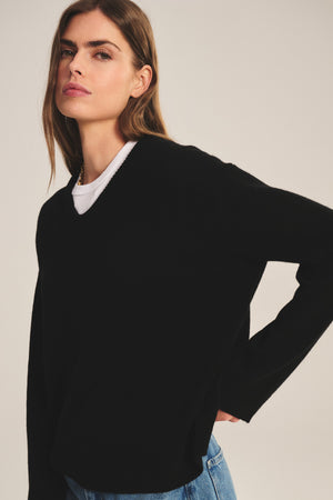 A woman with long brown hair wears the SORREL SWEATER from Velvet by Graham & Spencer—a modern black cashmere-wool top—layered over a white shirt and blue jeans, standing against a neutral background.