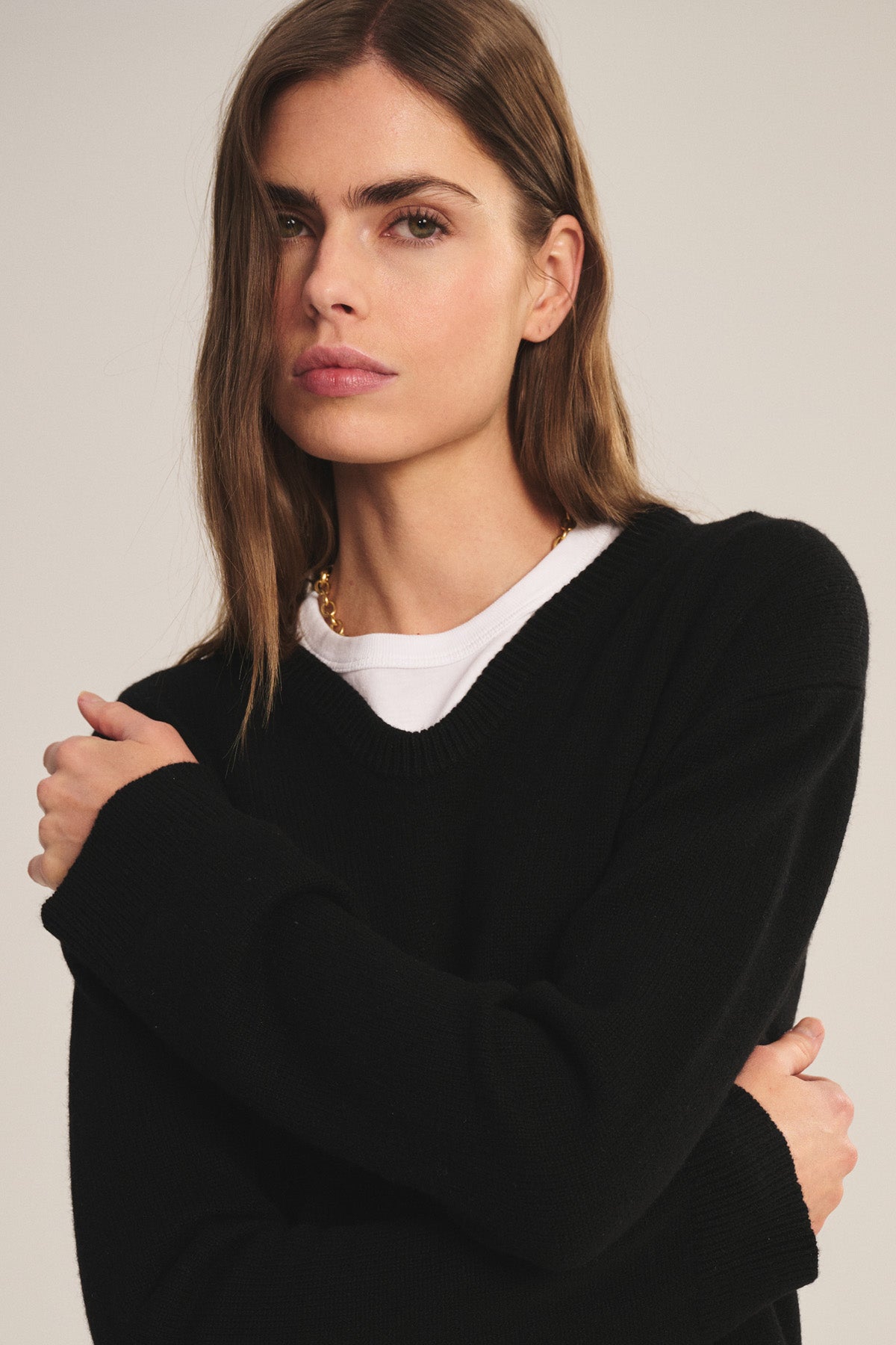 A woman with long brown hair stands with arms crossed, looking at the camera against a neutral background, wearing the SORREL SWEATER by Velvet by Graham & Spencer—a modern cashmere-wool blend over a white top.