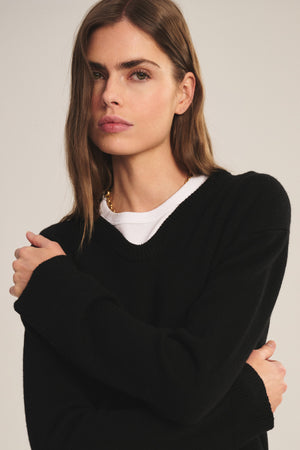 A woman with long brown hair stands with arms crossed, looking at the camera against a neutral background, wearing the SORREL SWEATER by Velvet by Graham & Spencer—a modern cashmere-wool blend over a white top.
