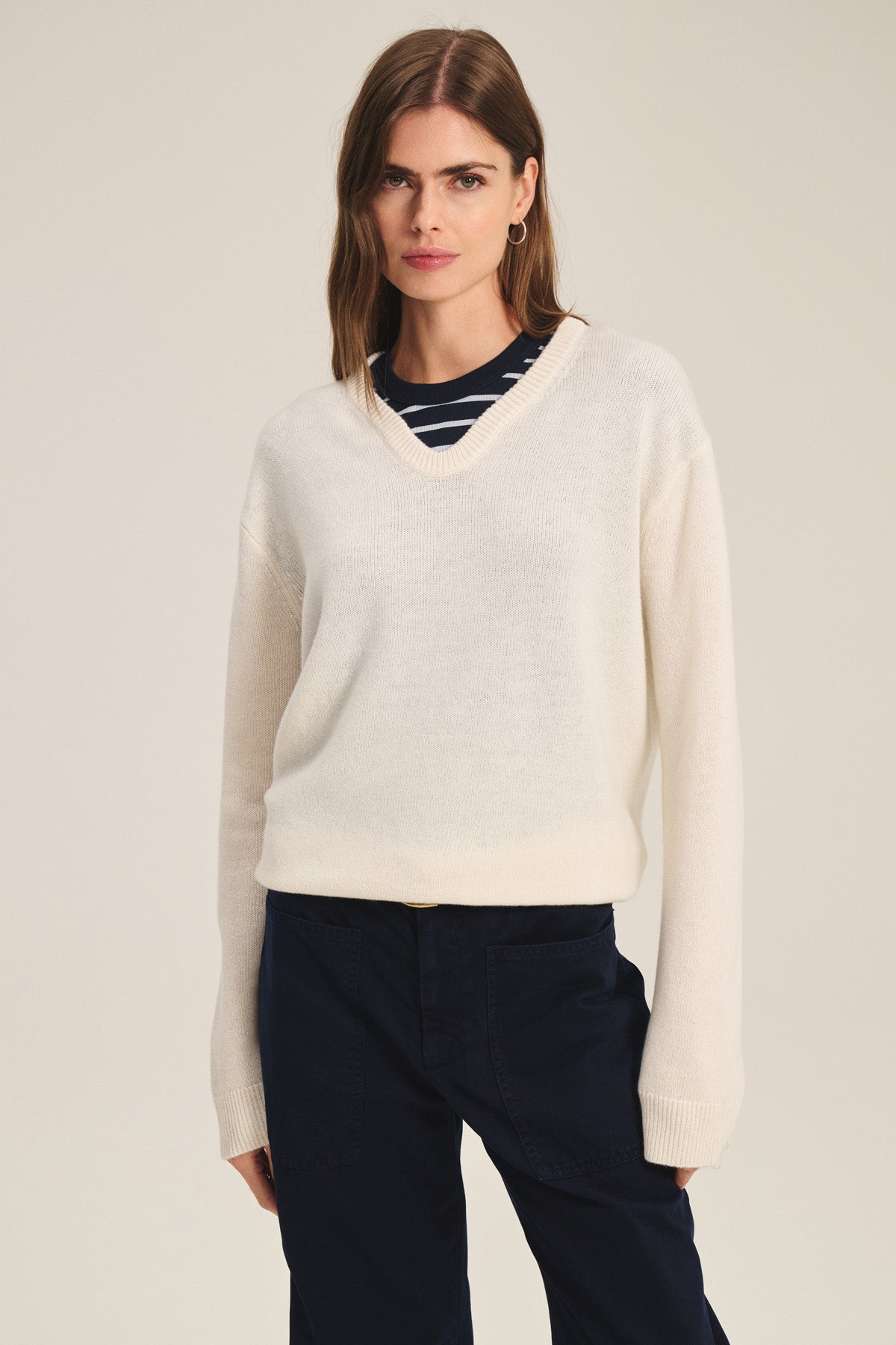 A woman wears the SORREL SWEATER by Velvet by Graham & Spencer, a cream cashmere-wool knit with a U-shaped neckline, layered over a navy and white striped top and paired with dark trousers, standing against a neutral background.-41436029747393
