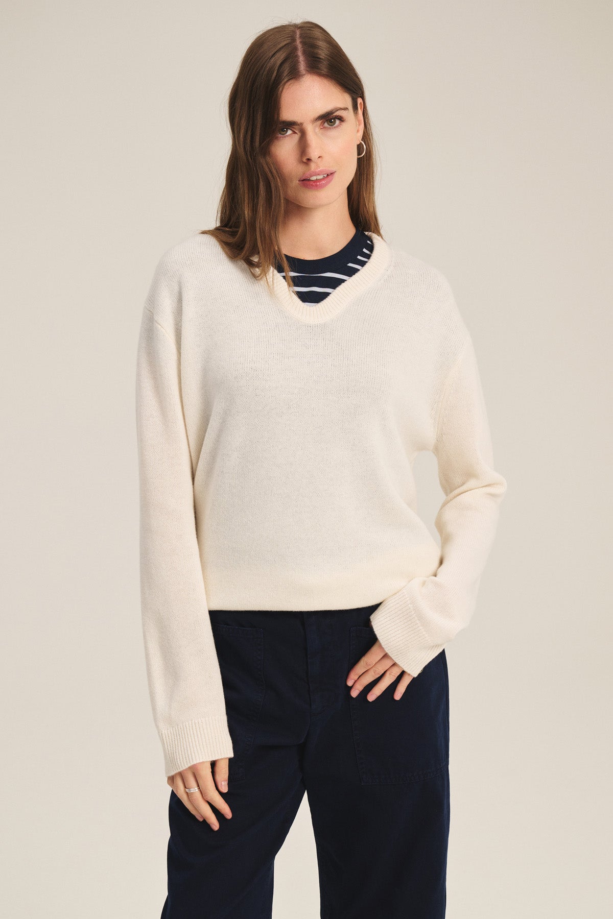 A woman wears the SORREL SWEATER by Velvet by Graham & Spencer, a cream cashmere-wool knit with a U-shaped neckline, layered over a navy and white striped top and dark pants, standing against a plain light background.-41436029780161