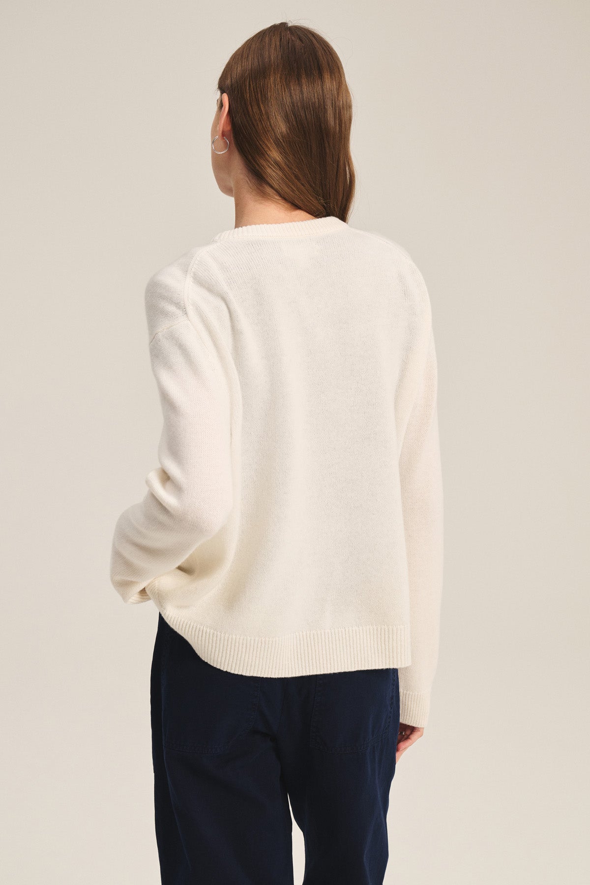 A person with long brown hair, wearing the Velvet by Graham & Spencer SORREL SWEATER—a cream cashmere-wool knit with a U-neckline—and dark pants stands facing away against a plain background.