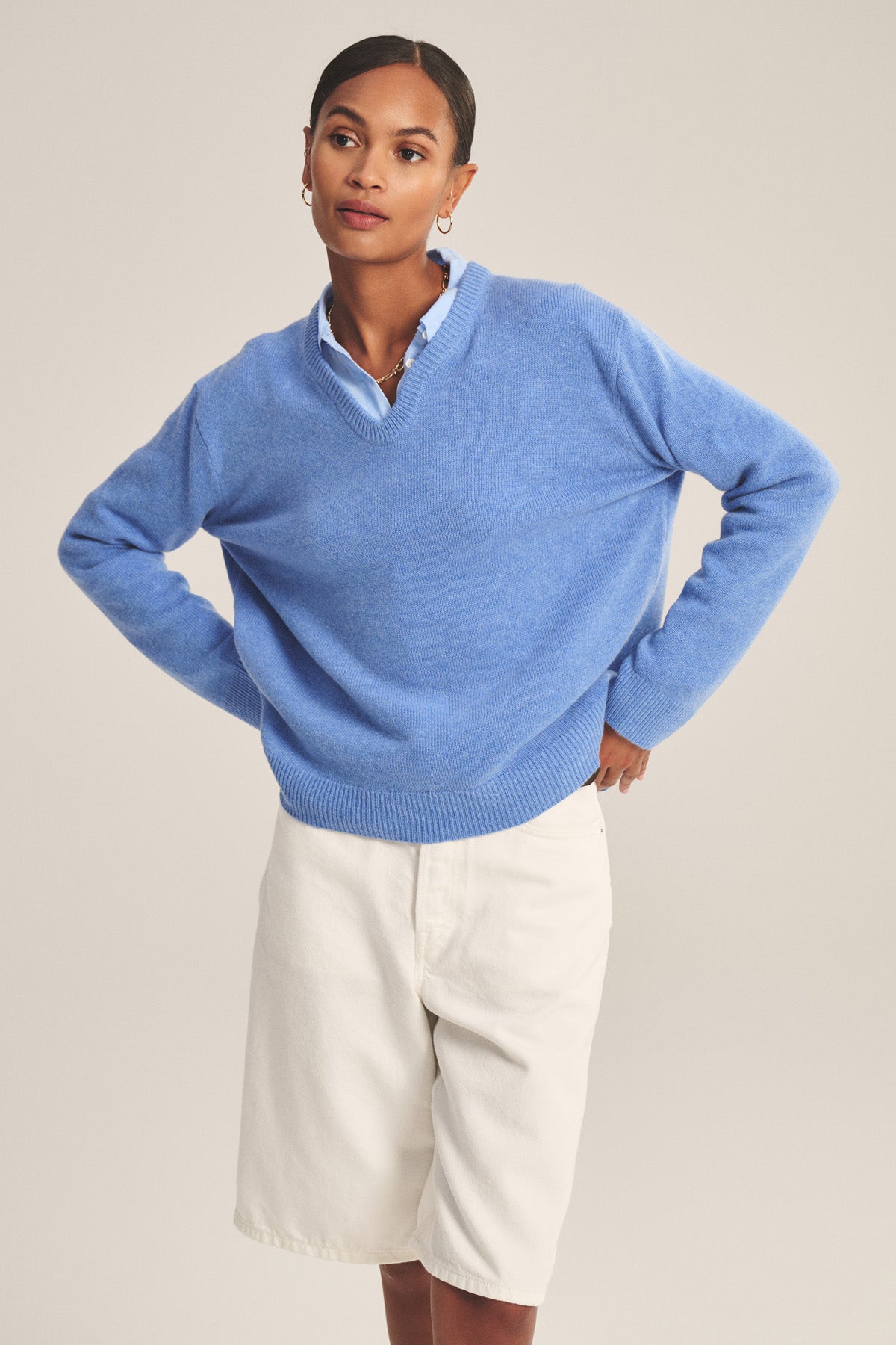 A person stands with hands on hips, wearing the SORREL SWEATER by Velvet by Graham & Spencer—a light blue cashmere-wool blend with a modern silhouette—layered over a collared shirt and paired with white knee-length shorts.-41008414949569