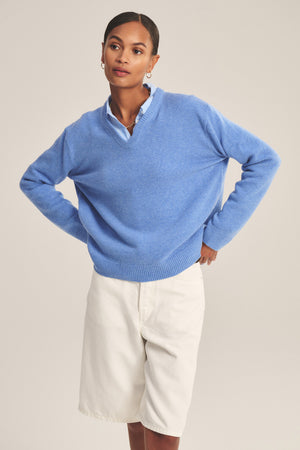 A person stands with hands on hips, wearing the SORREL SWEATER by Velvet by Graham & Spencer—a light blue cashmere-wool blend with a modern silhouette—layered over a collared shirt and paired with white knee-length shorts.