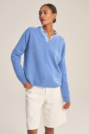 A woman stands against a plain background wearing the SORREL SWEATER by Velvet by Graham & Spencer—a light blue cashmere-wool piece with a modern silhouette—styled over a collared shirt and white knee-length shorts.