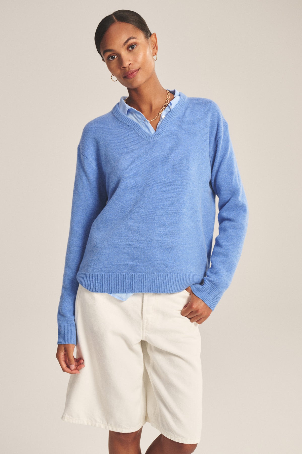 A person faces forward against a neutral background, wearing the SORREL SWEATER by Velvet by Graham & Spencer—a light blue cashmere-wool piece with a modern silhouette—over a collared shirt and white knee-length shorts.