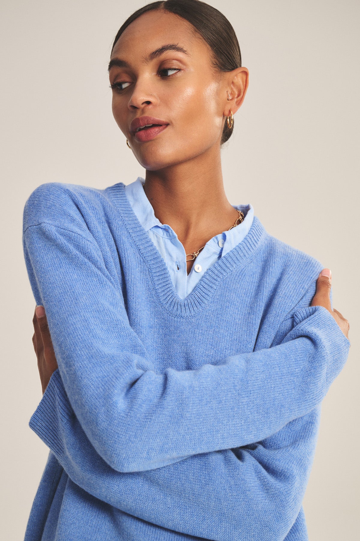 A woman with slicked-back hair wears the SORREL SWEATER by Velvet by Graham & Spencer, layered over a light blue collared shirt. She looks to the side with arms crossed, showcasing the sweater’s modern silhouette.
