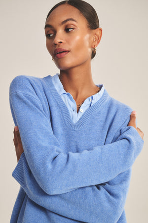 A woman with slicked-back hair wears the SORREL SWEATER by Velvet by Graham & Spencer, layered over a light blue collared shirt. She looks to the side with arms crossed, showcasing the sweater’s modern silhouette.