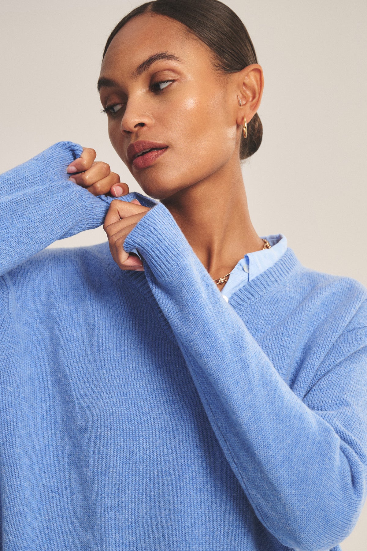 A woman wearing the SORREL SWEATER by Velvet by Graham & Spencer—a light blue cashmere-wool knit with a modern silhouette—adjusts her sleeve over a collared shirt, looking to the side.