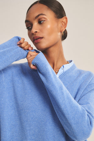 A woman wearing the SORREL SWEATER by Velvet by Graham & Spencer—a light blue cashmere-wool knit with a modern silhouette—adjusts her sleeve over a collared shirt, looking to the side.
