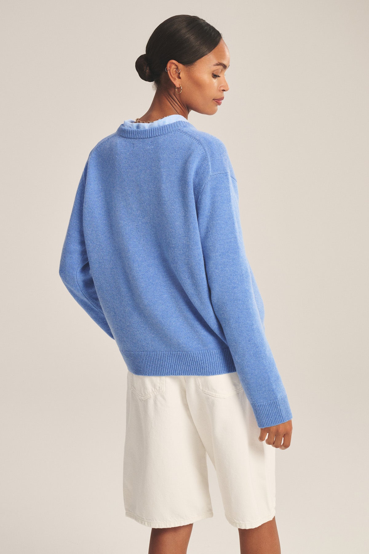 A woman faces away, wearing the SORREL SWEATER by Velvet by Graham & Spencer—a loose, light blue cashmere-wool sweater with a modern silhouette—paired with white knee-length shorts.-41008414752961