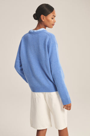 A woman faces away, wearing the SORREL SWEATER by Velvet by Graham & Spencer—a loose, light blue cashmere-wool sweater with a modern silhouette—paired with white knee-length shorts.