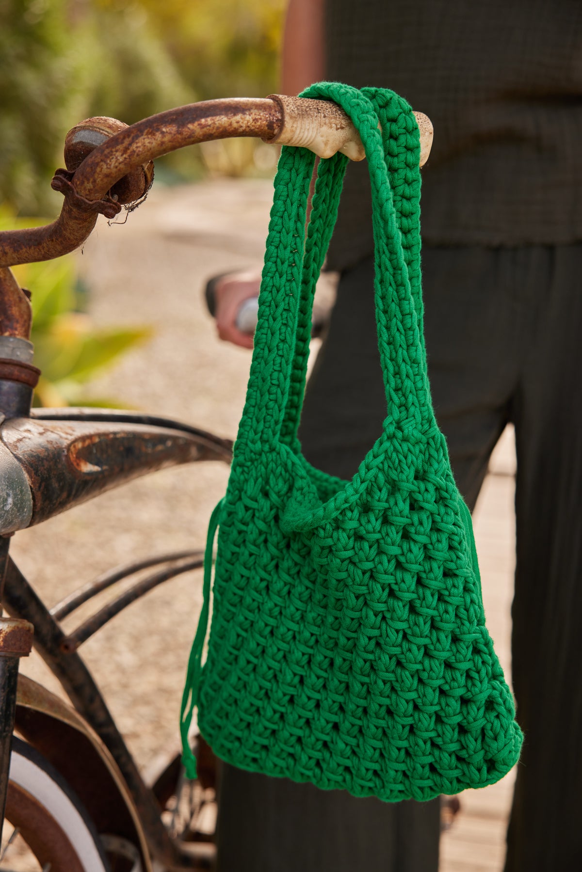   A green, knitted PENNY CROCHET BAG by Velvet by Jenny Graham with a drawstring lining hangs on the handlebar of a vintage bicycle. A person dressed in dark clothing stands in the background. 