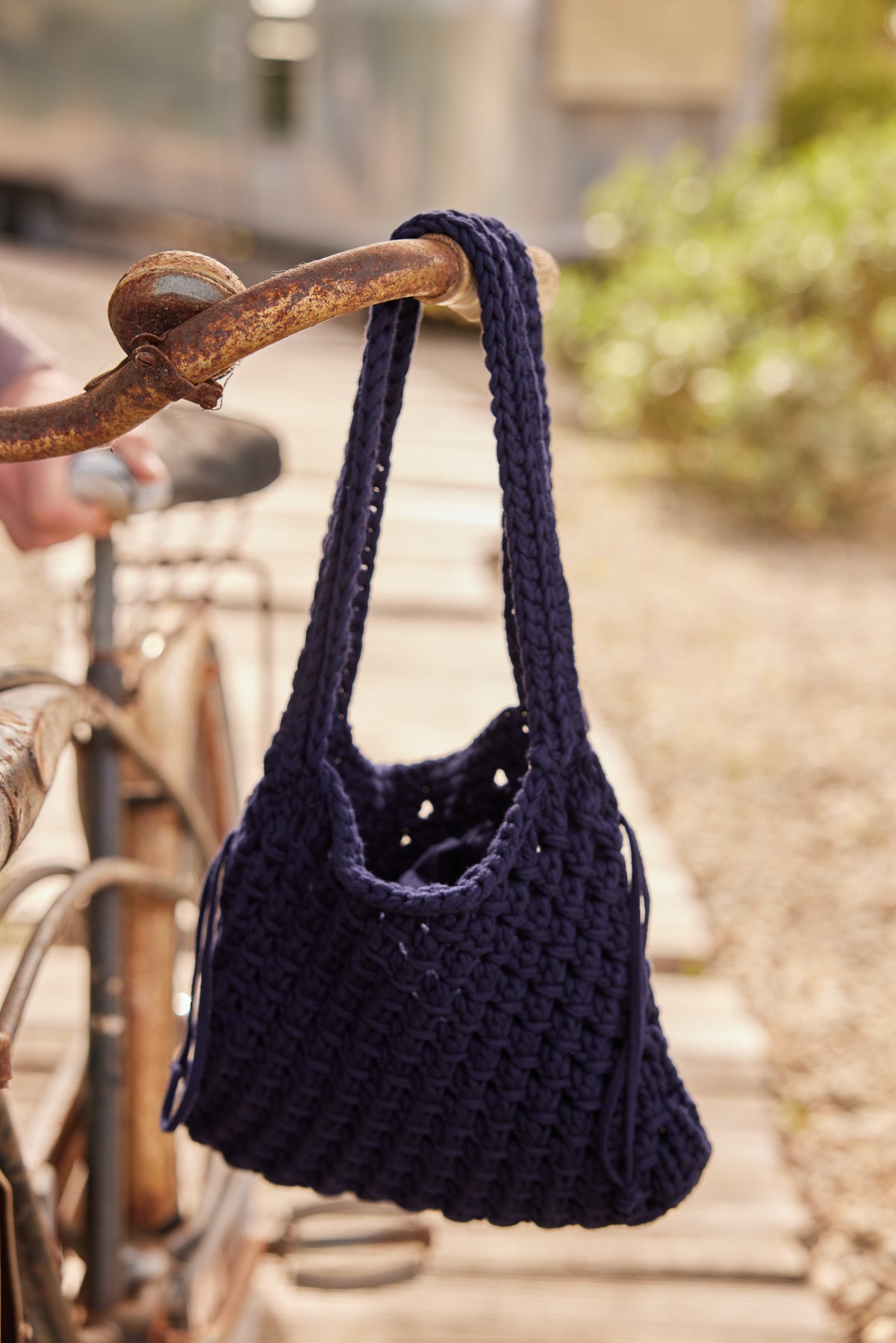   A PENNY CROCHET BAG by Velvet by Jenny Graham, crafted with chunky cotton cord and featuring a drawstring lining, hangs on the rusted handlebars of a vintage bicycle on a wooden path with greenery in the background. 