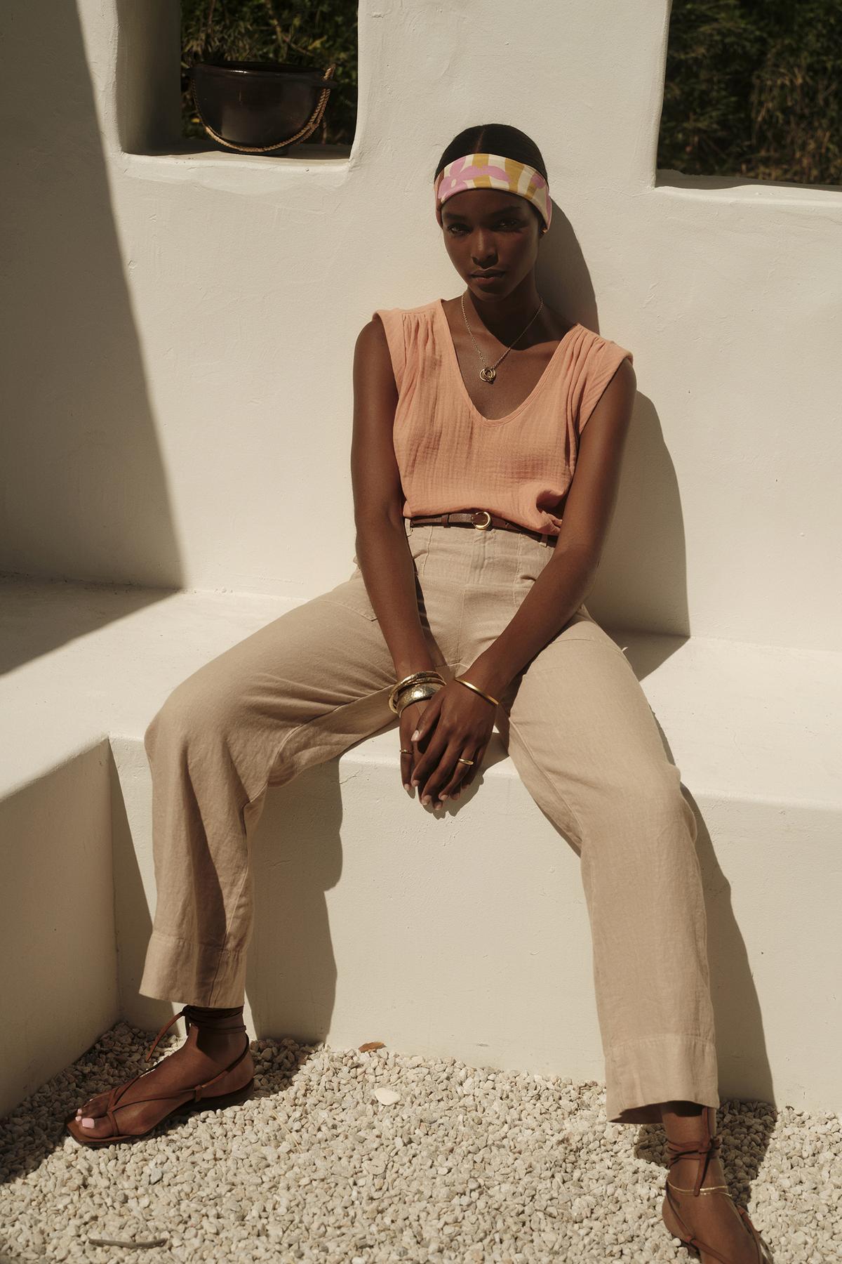   A person wearing the JAYLA COTTON GAUZE TANK TOP by Velvet by Graham & Spencer, along with beige trousers and sandals, sits on a white bench against a white wall. The individual accessorizes with a colorful headband, bracelets, and a necklace. 