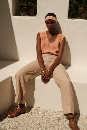 A person wearing the JAYLA COTTON GAUZE TANK TOP by Velvet by Graham & Spencer, along with beige trousers and sandals, sits on a white bench against a white wall. The individual accessorizes with a colorful headband, bracelets, and a necklace.