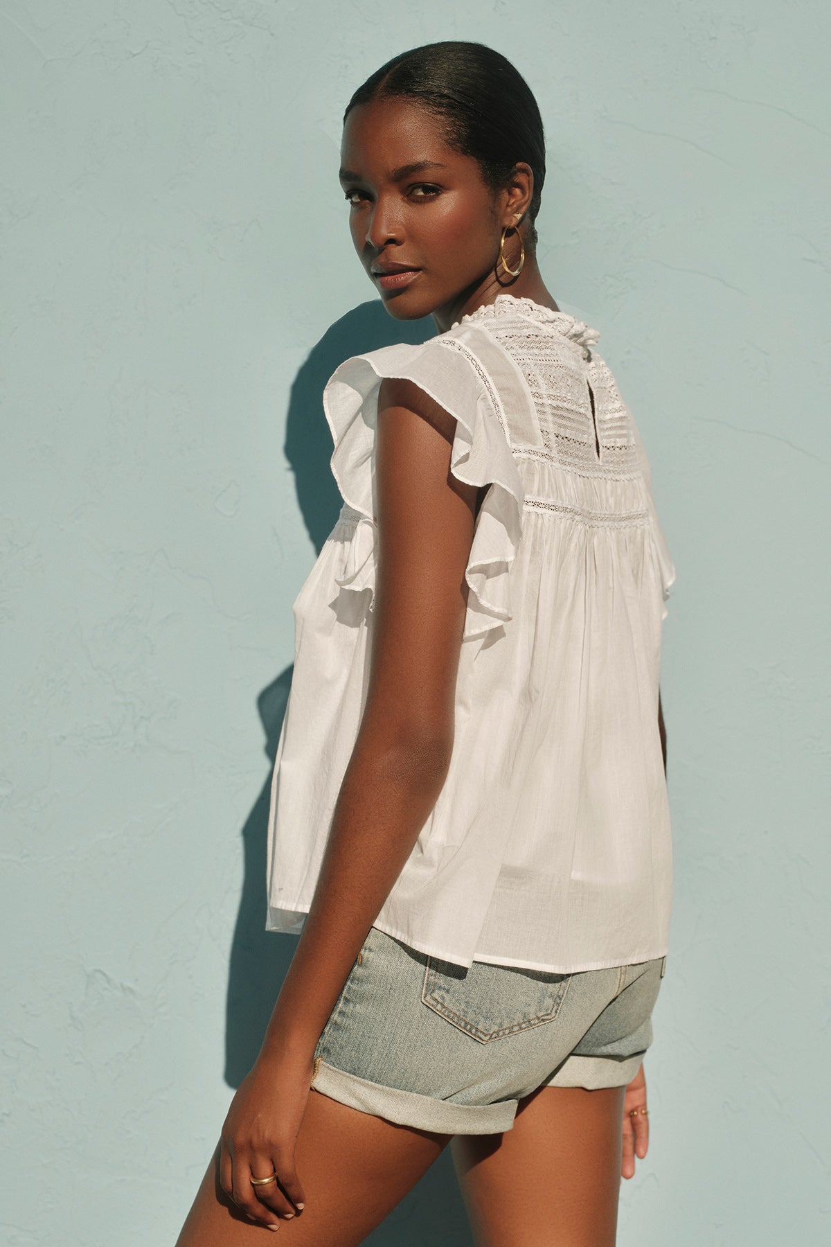   A person in the INESSA COTTON LACE TOP by Velvet by Graham & Spencer and denim shorts stands against a light blue textured wall, looking over their shoulder. 