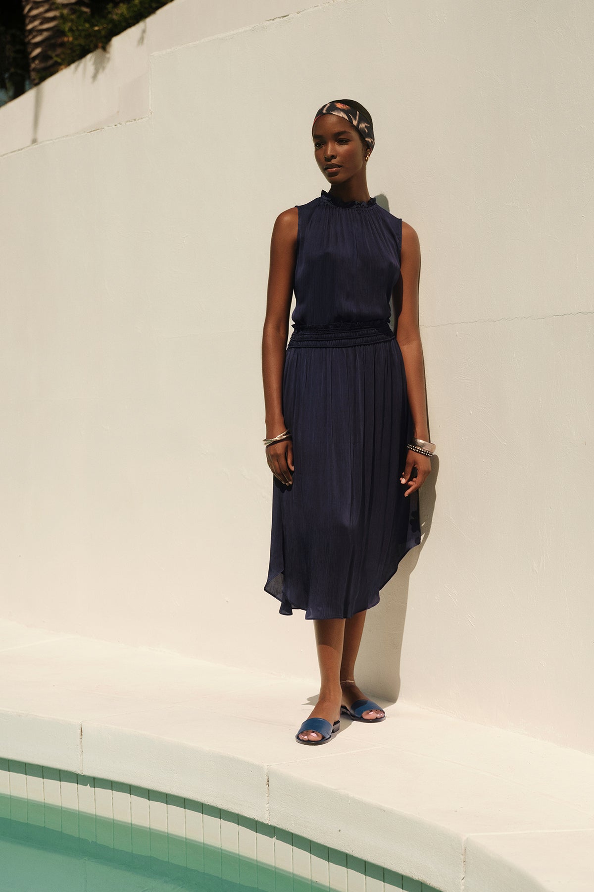 A woman in a sleeveless navy dress with DIMI SMOCKED SKIRT detailing standing beside a pool, with her shadow casting on the wall.-36409579897025