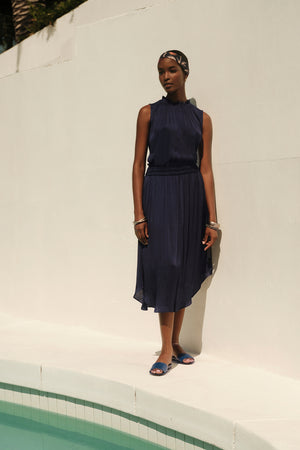 A woman in a KIANA RUFFLE NECK TANK TOP by Velvet by Graham & Spencer and headscarf stands beside a swimming pool, sunlight casting shadows on a white wall behind her.