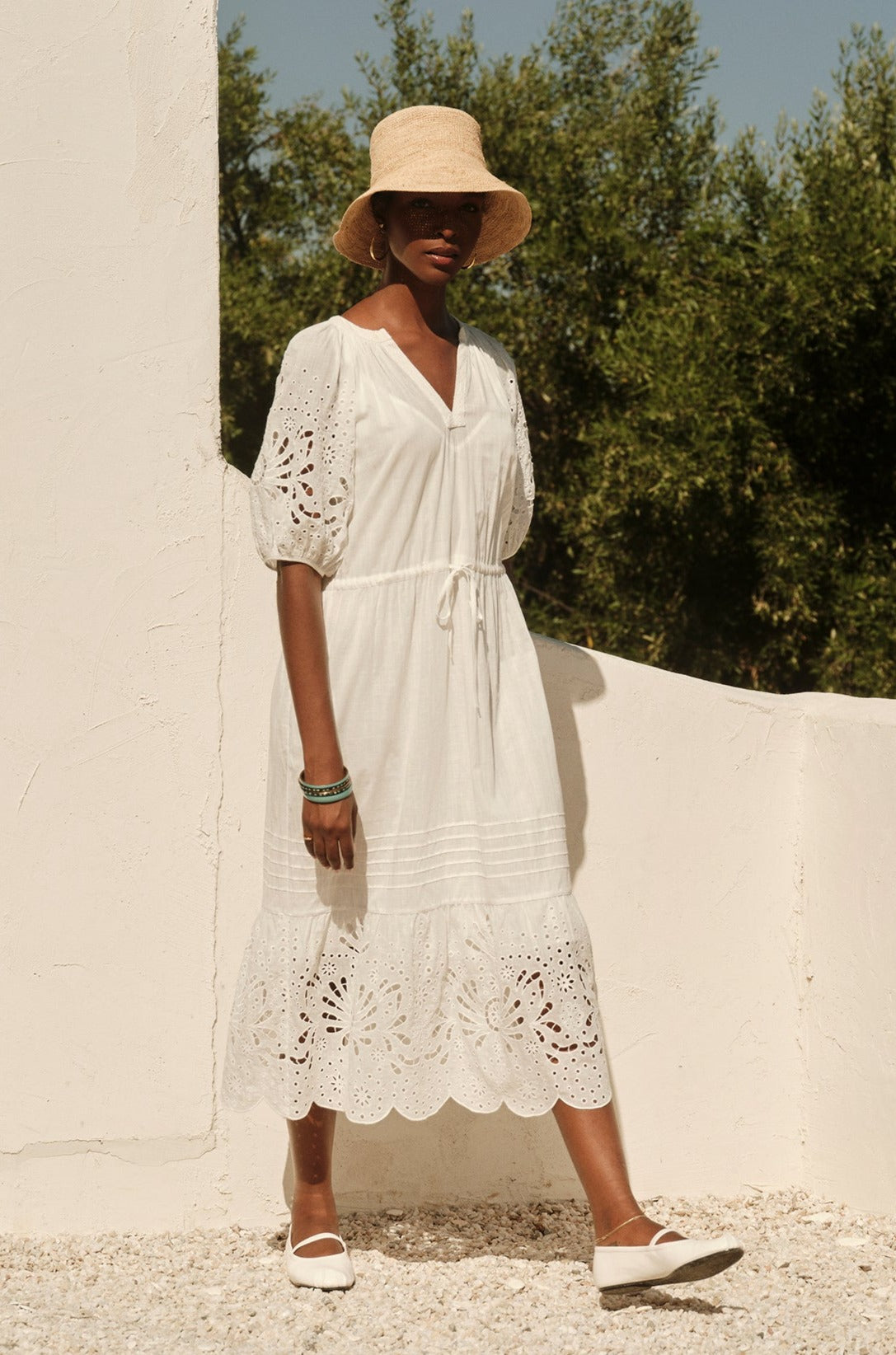 A woman in a Velvet by Graham & Spencer NADIA EMBROIDERED COTTON LACE DRESS and straw hat stands against a white wall, surrounded by gravel and greenery, on a sunny day.-36483131080897