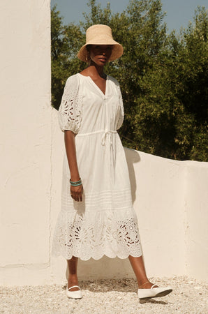 A woman in a Velvet by Graham & Spencer NADIA EMBROIDERED COTTON LACE DRESS and straw hat stands against a white wall, surrounded by gravel and greenery, on a sunny day.