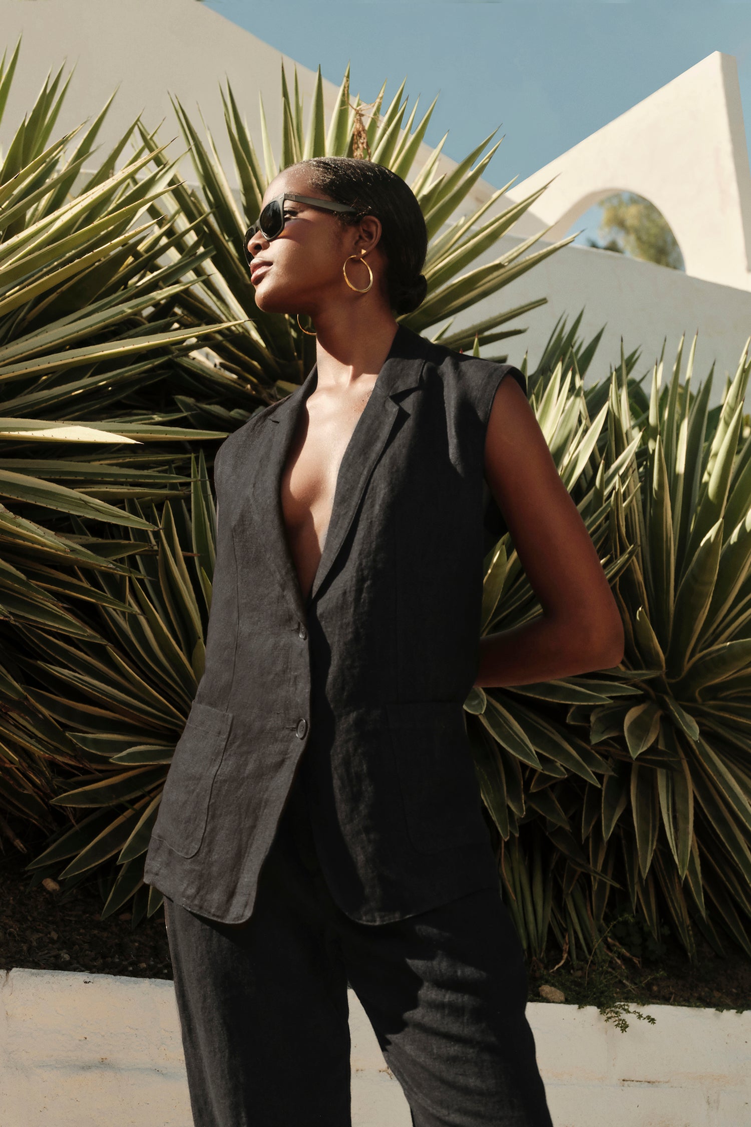   A stylish woman in a black BETHAN HEAVY LINEN SLEEVELESS BLAZER by Velvet by Graham & Spencer stands confidently against a background of sharp green leaves, wearing sunglasses and hoop earrings. 