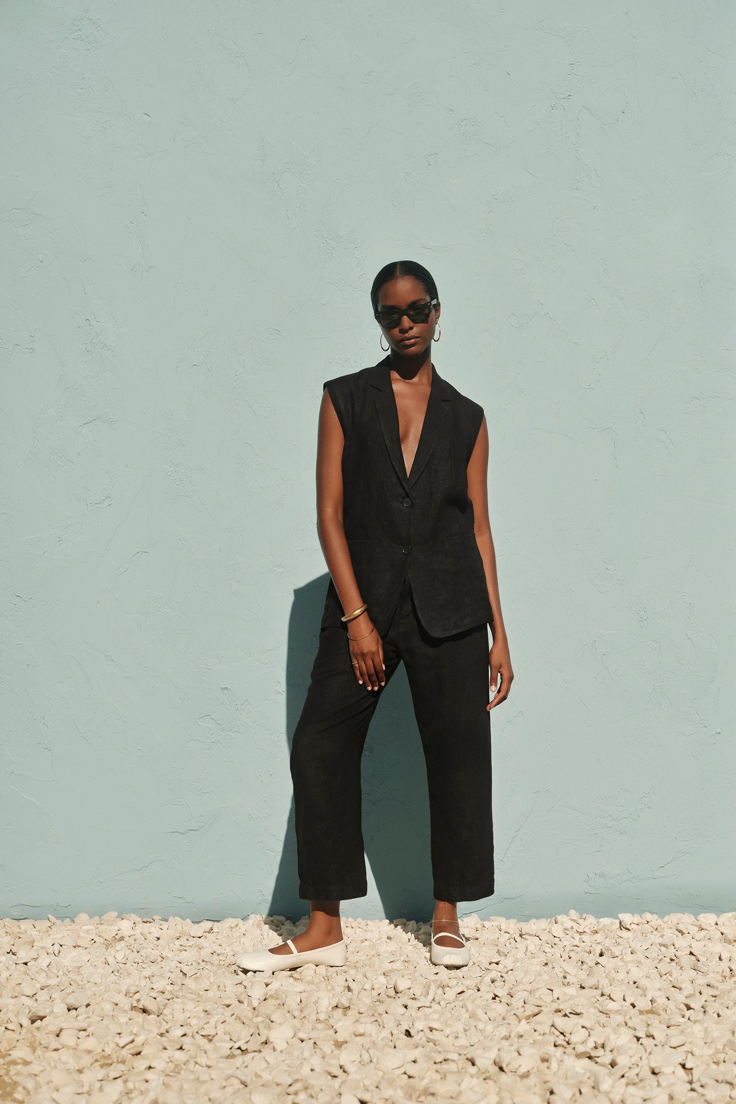   A woman stands confidently against a light blue wall, wearing a Velvet by Graham & Spencer BETHAN HEAVY LINEN SLEEVELESS BLAZER and sunglasses, with her hands in pockets. 