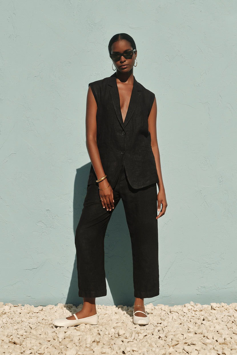 A woman stands against a light blue wall, dressed in a black sleeveless vest and Velvet by Graham & Spencer's JESSIE HEAVY LINEN PANT, accessorized with sunglasses and white shoes.