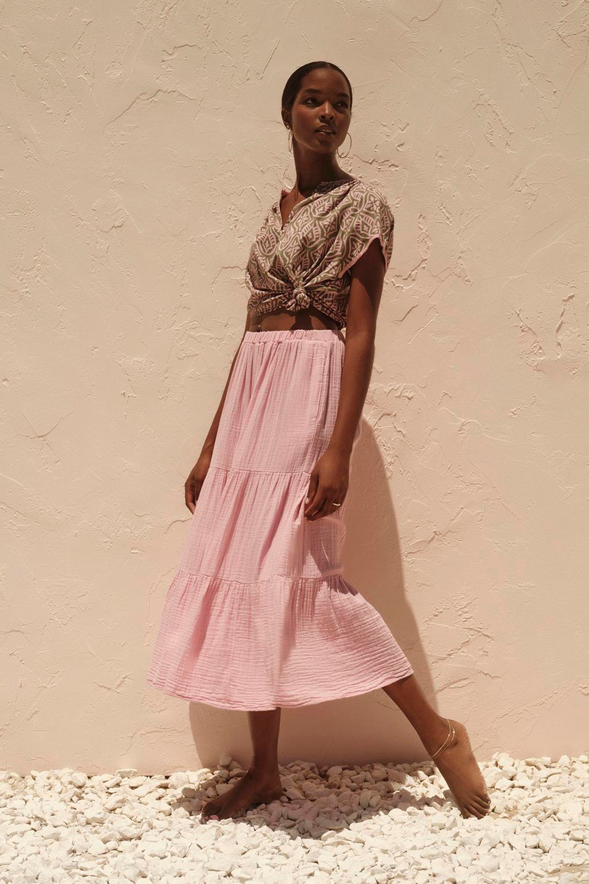   A woman in a pink Danielle Cotton Gauze Tiered Skirt by Velvet by Graham & Spencer with in-seam pockets standing next to a cotton gauze wall. 