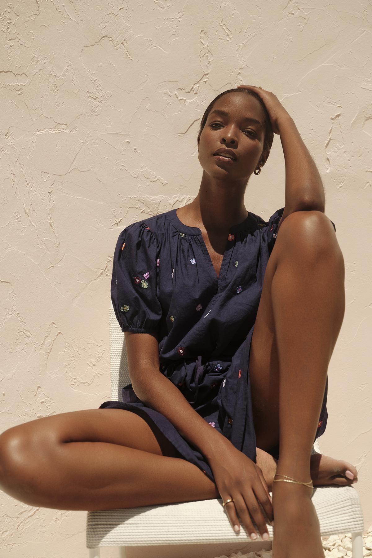 A woman in a CLEO EMBROIDERED BOHO DRESS by Velvet by Graham & Spencer with a v-neckline sits on a white bench, hand on head, against a textured cream wall, in sunlight.-36320306069697