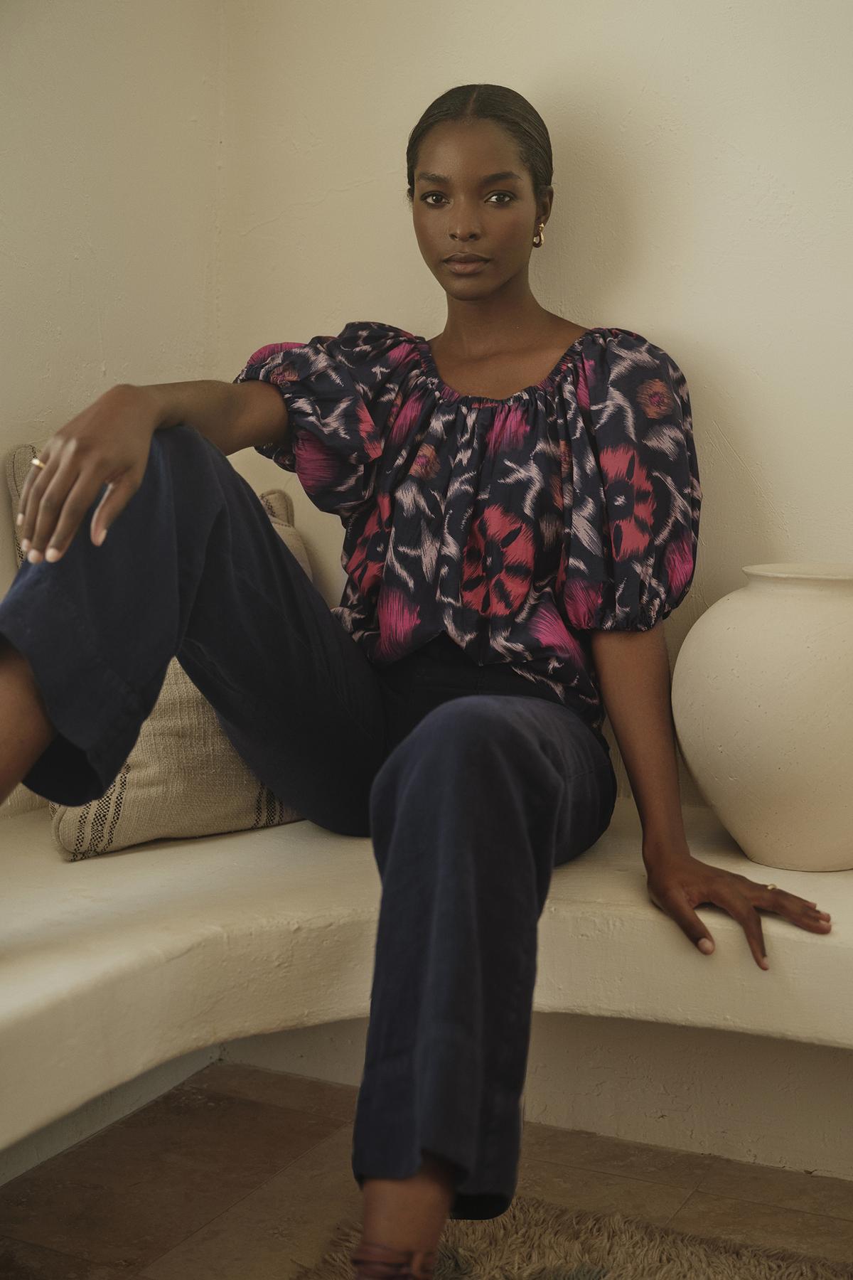 A woman sits casually on a bench with a neutral expression, wearing a patterned blouse and dark, wide-leg Velvet by Graham & Spencer DRU HEAVY LINEN PANT.-36320310493377