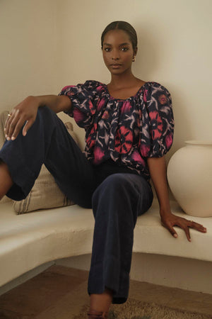 A woman sits casually on a bench with a neutral expression, wearing a patterned blouse and dark, wide-leg Velvet by Graham & Spencer DRU HEAVY LINEN PANT.