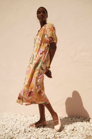 A woman in a flowing, pastel-colored CAROL PRINTED BOHO DRESS by Velvet by Graham & Spencer with a detachable twist braid belt stands against a light beige wall, casting a long shadow on a white pebbled surface.