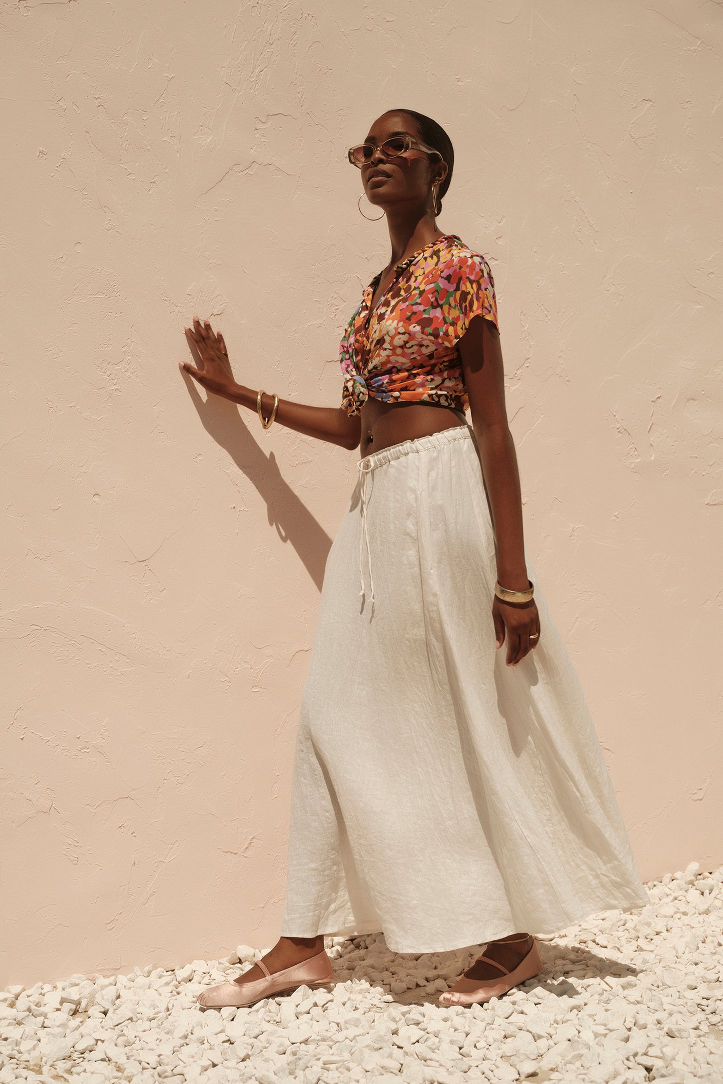 A woman in sunglasses, a floral crop top, and the Velvet by Graham & Spencer BAILEY LINEN MAXI SKIRT stands confidently against a pale textured wall.-36691390267585
