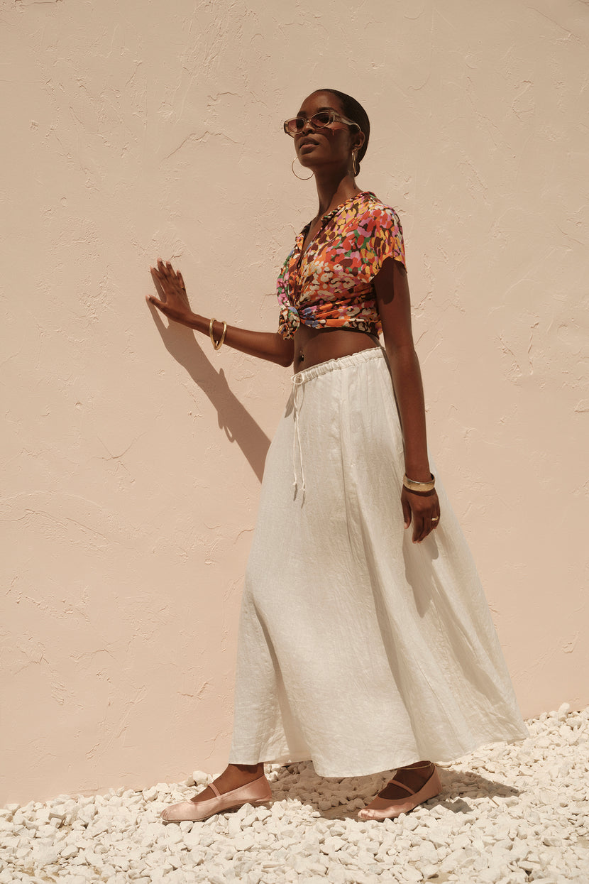 A woman in sunglasses, a floral crop top, and the Velvet by Graham & Spencer BAILEY LINEN MAXI SKIRT stands confidently against a pale textured wall.